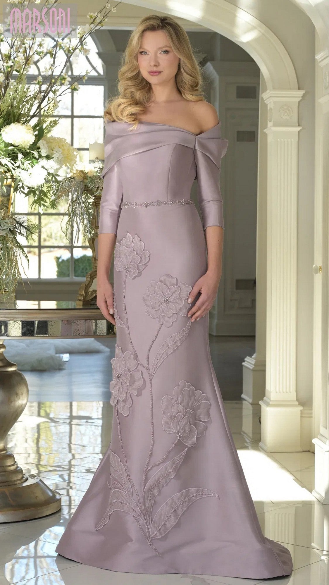 A woman stands indoors wearing the Marsoni MV1374 long formal dress in dusty rose, featuring a fitted, off-shoulder design with floral appliqué details and a thin jeweled belt.
